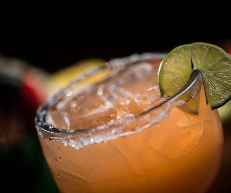 Orange cocktail with ice, a salted rim, and a lime wedge garnish.