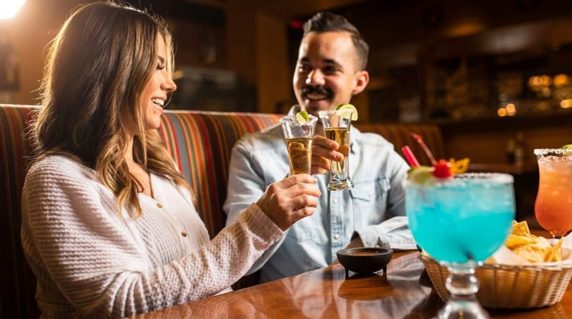 Smiling couple toasting with tequila shots at a vibrant restaurant table with colorful drinks.